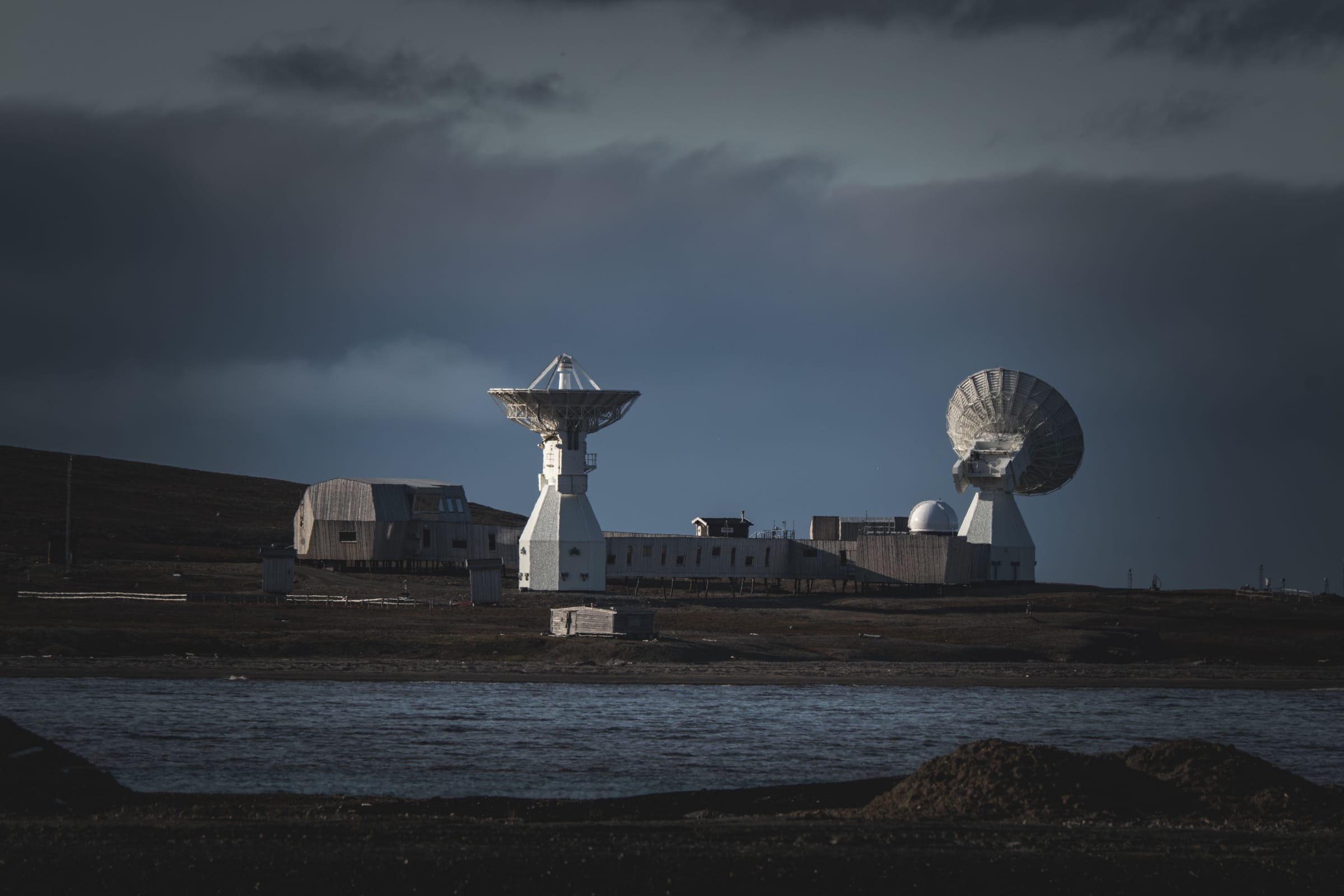 Two large satellite dishes and low buildings at a remote ground station beside water, under a dark cloudy sky.